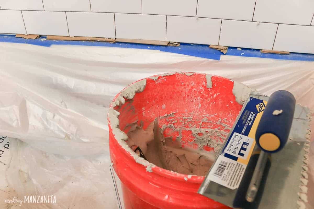 Bucket of thinset mortar sitting in a bathtub with a trowel sitting on it while white subway tile gets installed on shower walls
