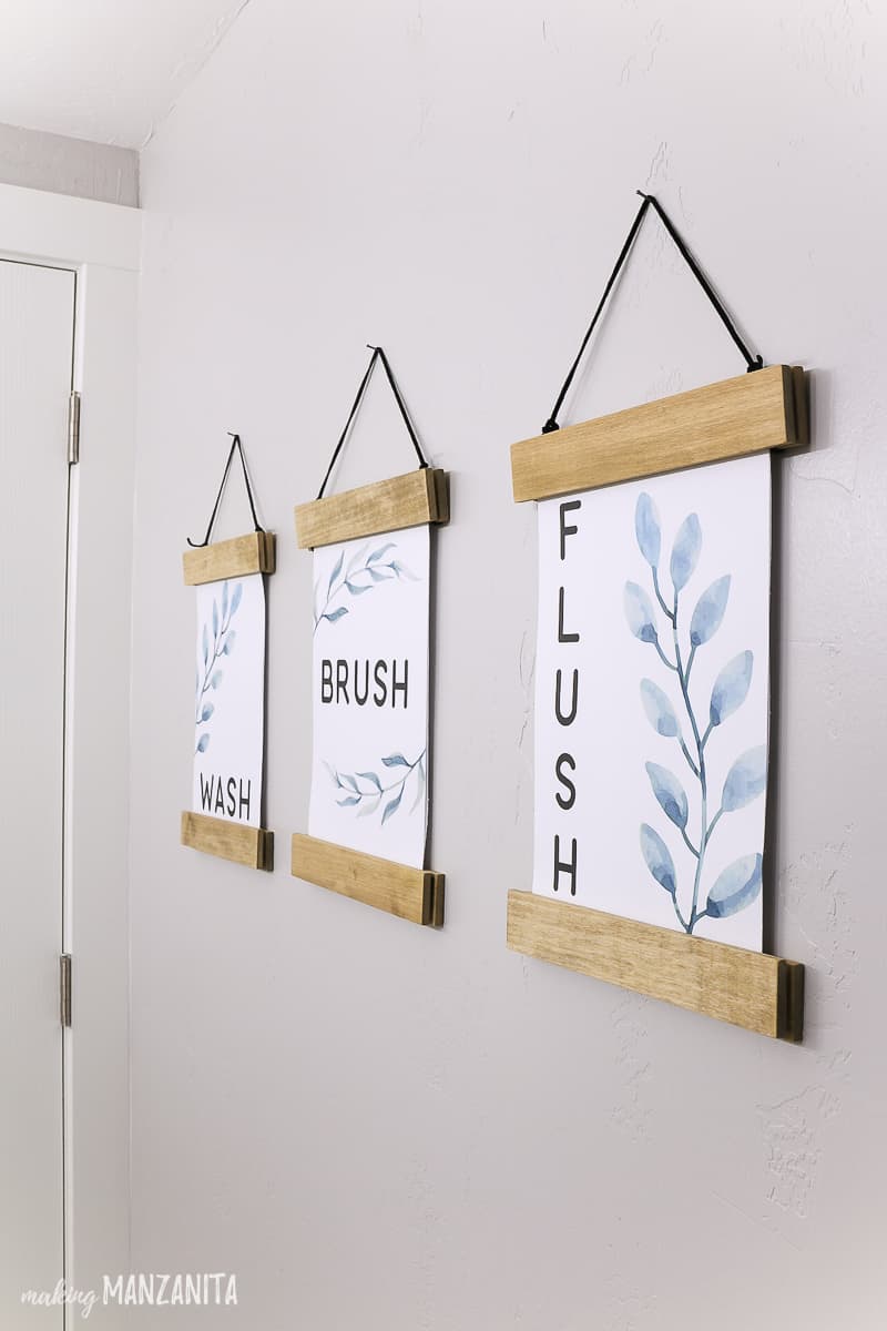 Three side by side wall decor printables with wash brush flush on them with leaves and floral designs hanging in DIY wood frames on a wall in a farmhouse bathroom