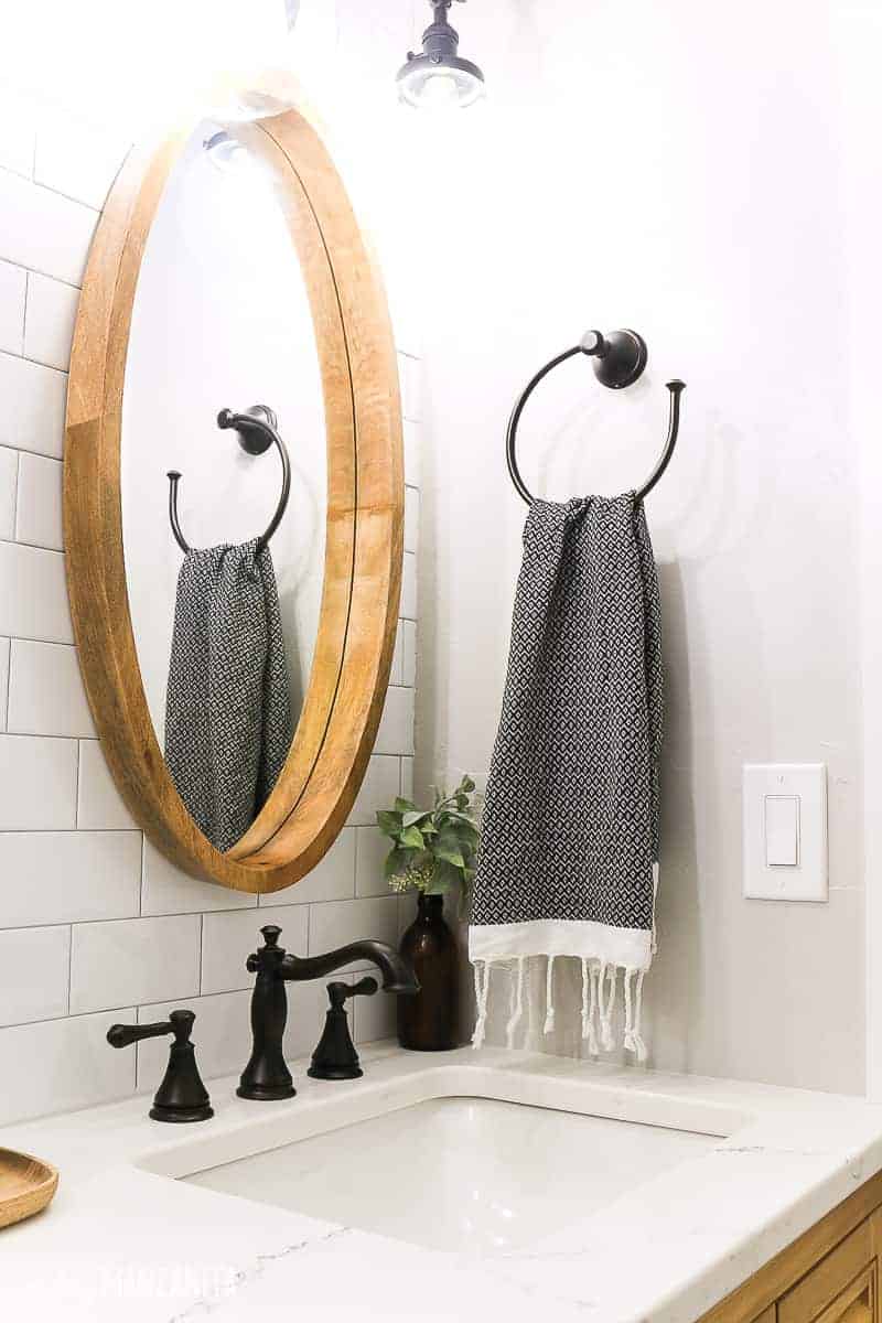 Wood framed oval mirror mounted on white subway tile wall over a vanity with oil rubbed bronze sink faucet and white quartz countertop with vanity light above it. Black turkish towel hanging on towel ring on wall