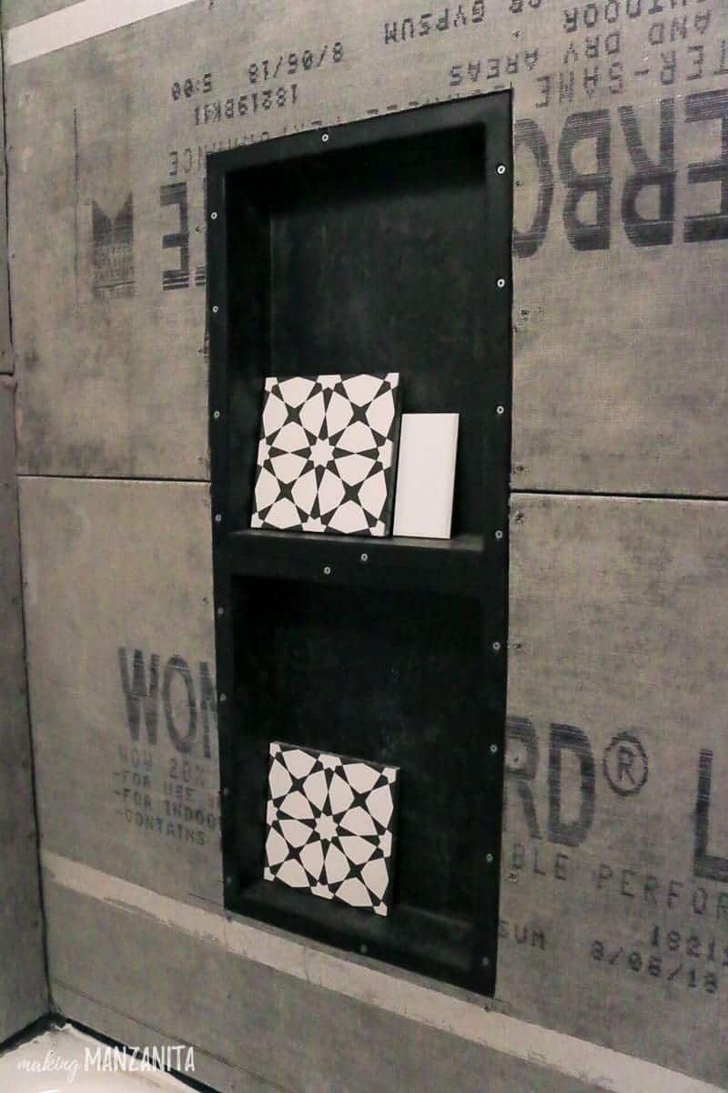 The process of installing a shower niche during bathroom renovation; a prefabricated black shower niche installed in the wall with black and white geometric patterned tile that will be installed in the shower niche backsplash.