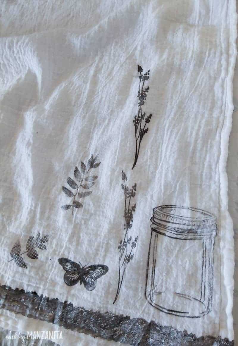 A close up image of the corner of a white linen tea towel, stamped with black fabric paint designs of a mason jar, springs of leaves and flowers, and a small butterlfy.