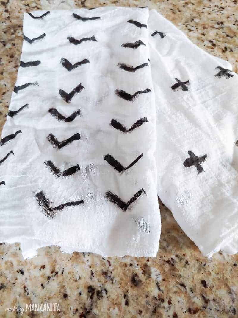 Two white fabric tea towels lay on a granite countertop. Each towel is stamped with a custom design in black fabric paint -- one towel has rows of black arrows, and the other has swiss crosses across the towel.