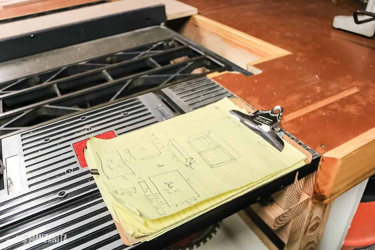 A note pad on a clip board sits on a work bench, detailing the build plans for a hallway cabinet.