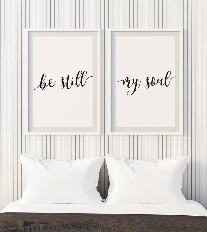 Looking for above bed decor in master bedroom? Try white pillows and white vertical shiplap wall behind bed with two framed printables hanging that say be still and my soul in black script lettering