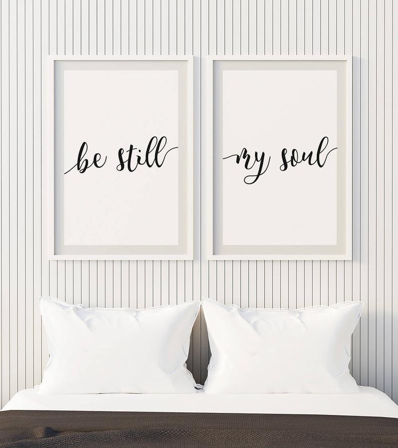 Looking for above bed decor in master bedroom? Try white pillows and white vertical shiplap wall behind bed with two framed printables hanging that say be still and my soul in black script lettering 