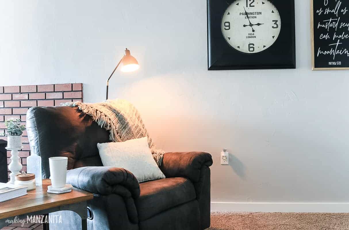 Black modern farmhouse floor lamp behind recliner with blanket draped on back and white throw pillow in cozy living room with industrial clock on wall
