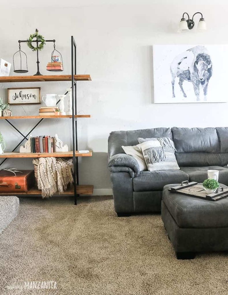 Farmhouse decorated living room with gray couch and ottoman with buffalo watercolor art on wall above couch and shelving unit next to couch decorated with farmhouse style vintage decor like old books, vintage sale, white farmhouse pitcher, candleholders and big wicker basket for blanket storage with light gray painted walls with Behr Chic gray paint color