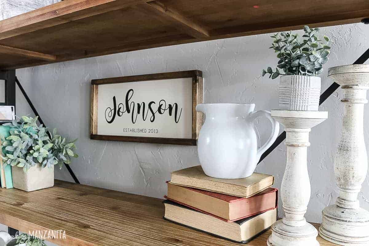 One shelf of our living room bookshelf, decorated with vintage books, candle holders, faux greenery and a farmhouse white ceramic pitcher.