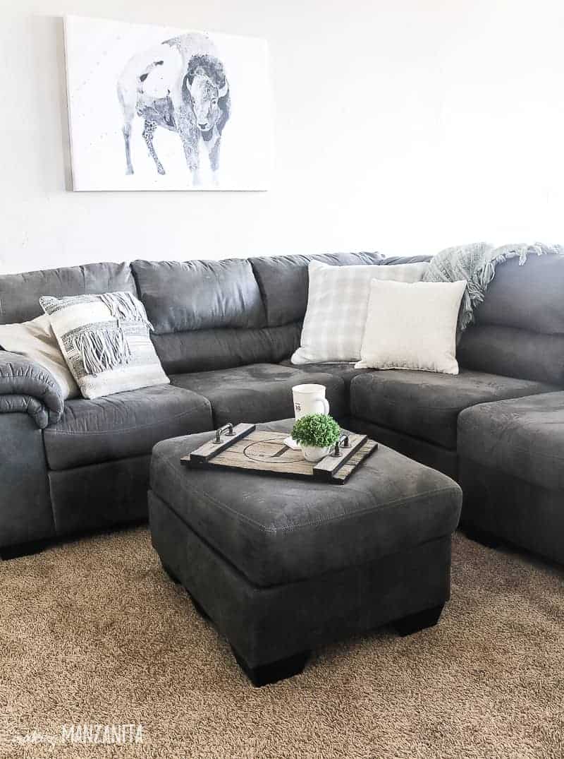 Corner of dark gray sectional couch with throw pillows with boho farmhouse style and matching gray ottoman with wood tray holding coffee cup and mini plant