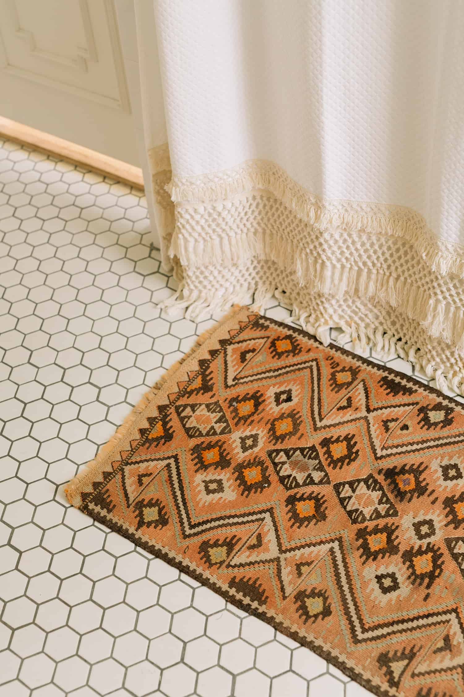 Tan southwestern looking vintage rug in a bathroom with white hexagon floor tile and macrame shower curtain