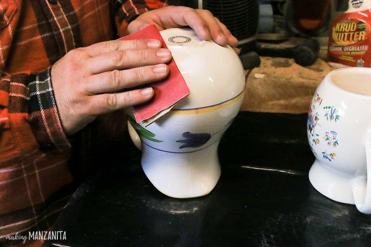 Using sandpaper to lightly sand the glaze finish off of a thrifted white ceramic pitcher.