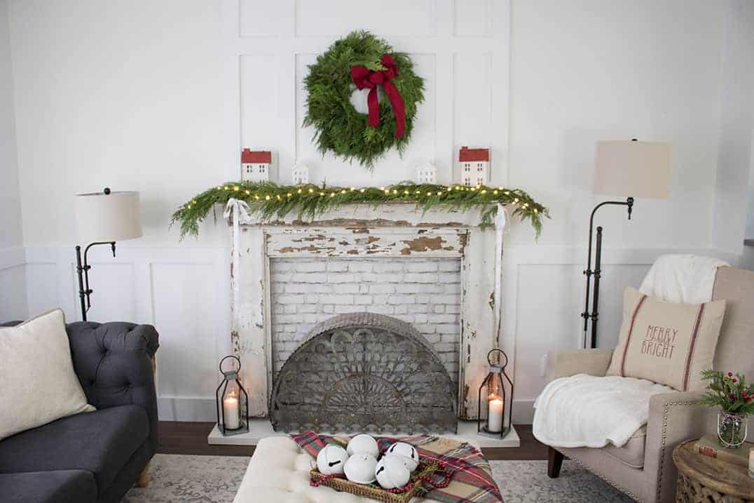 farmhouse decorated living room with christmas decorations featuring a vintage mantel with white chipped paint wood, greenery and lights and christmas wreath hanging above it. Two farmhouse floor lamps, a tan chair and dark gray couch are also shown in photo