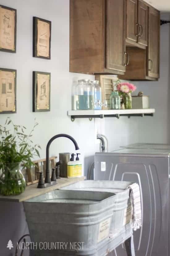 Galvanized tubs used as a sink in laundry room with vintage inspired wall decor, shelf holding laundry supplies, upper wood cabinets and washer and dryer shown in background