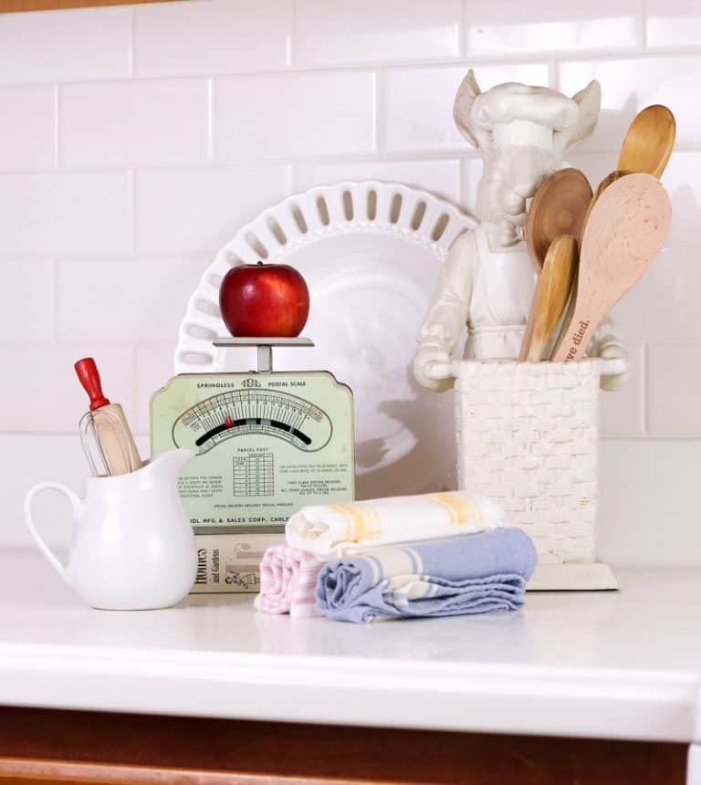 Vintage scale sitting on kitchen counter with red apple on it. Countertop also features white farmhouse pitcher, rolled hand towels, wooden spoons and white subway tile is shown in the background