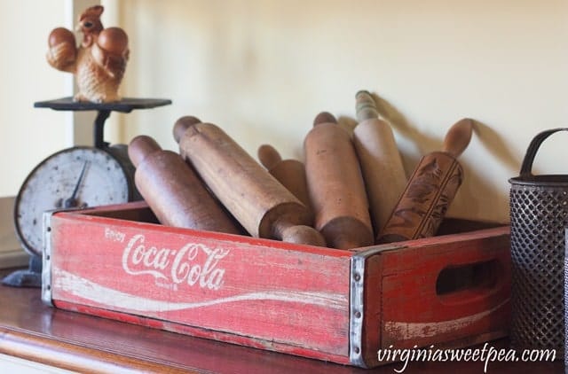 Old vintage coca-cola red crate filled with antique rolling pins in kitchen with vintage scale in the background
