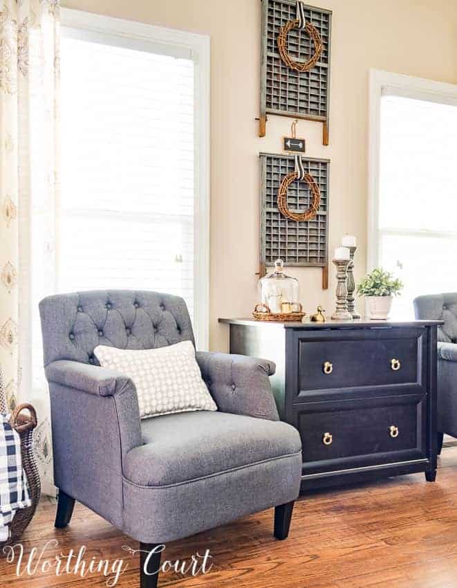 Vintage letterpress drawers hanging on wall with grapevine wreaths as wall decor with gray chair in front of window, tan pillow and black painted cabinet