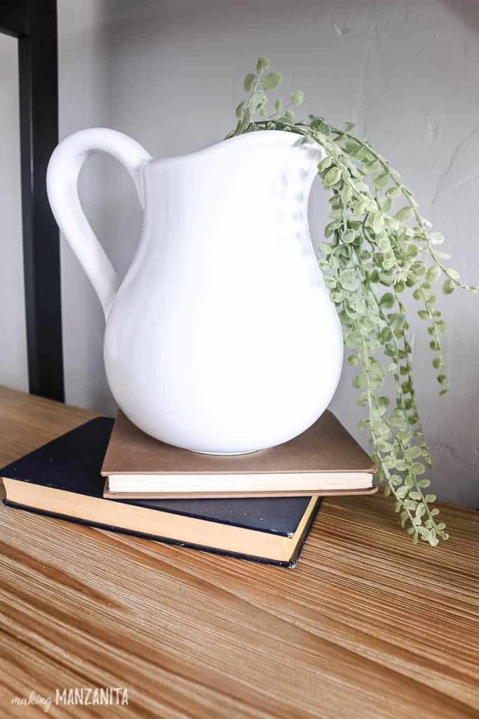A white farmhouse ceramic pitcher - upcycled from thrifted ceramic pitchers - sits on a shelf on a stack of books