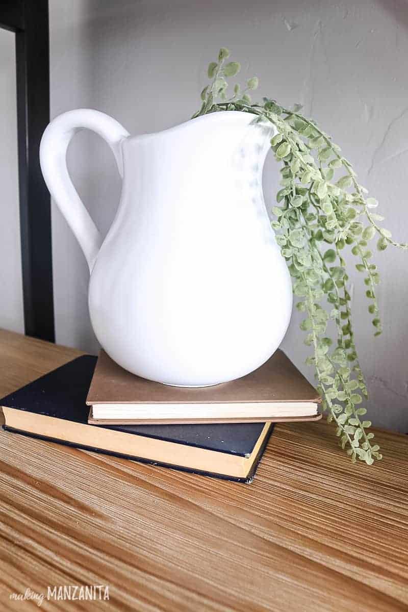 White farmhouse pitcher made by spray painting an old thrift store ceramic pitcher with a a high gloss white spray sitting on 2 vintage books stacked on a wooden shelf with faux greenery in it