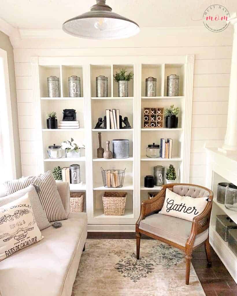 Built in shelves with vintage decor, books, baskets and farmhouse style decorations in a room with shiplap walls, industrial lighting and cream colored couch