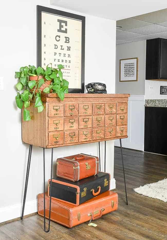 Vintage card catalog with hairpin legs restored and staged with old suitcases underneath, vintage eye chart as wall decor and plant and vintage telephone as decor