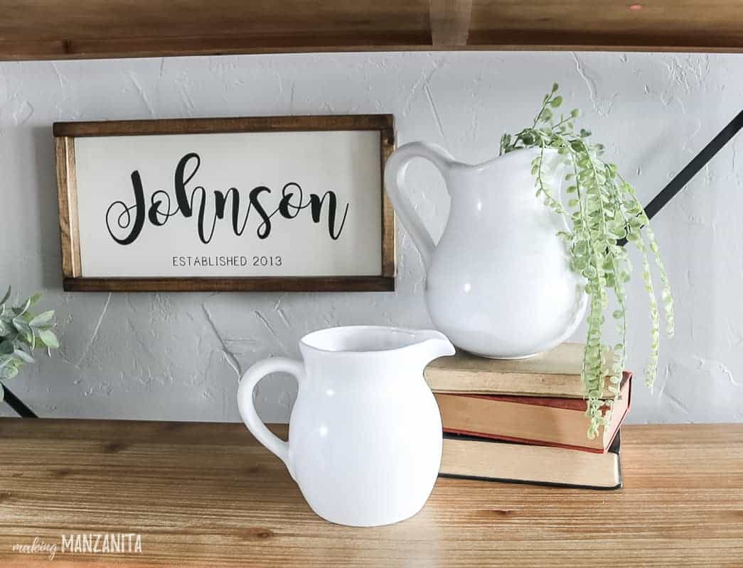 Shelf decor idea with a farmhouse sign hanging on the wall in the background that says Johnson and 2 white farmhouse pitchers displayed with a stack of vintage books and faux greenery