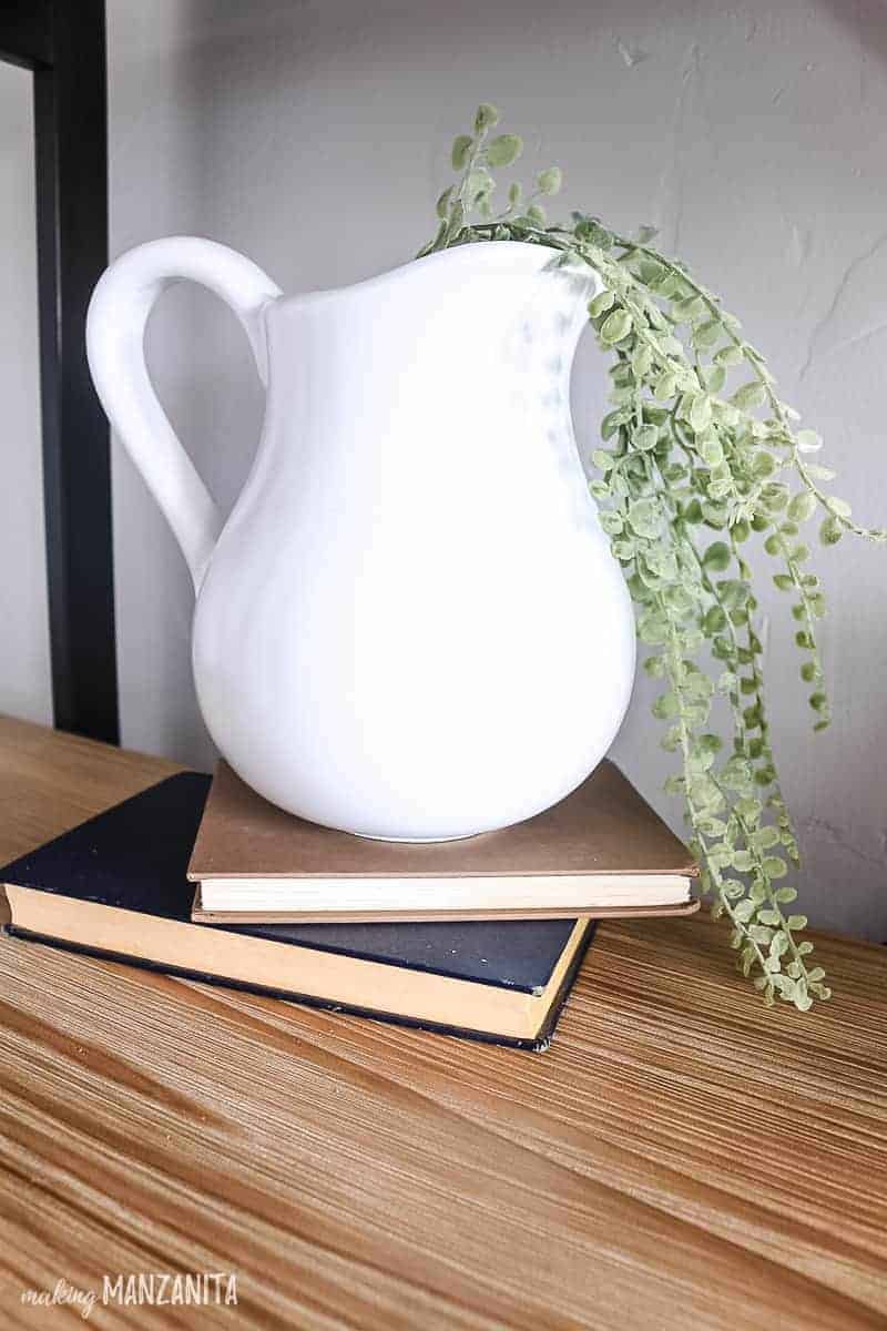 Vintage books on a wood shelf topped with a white pitcher with faux greenery flowing out of it