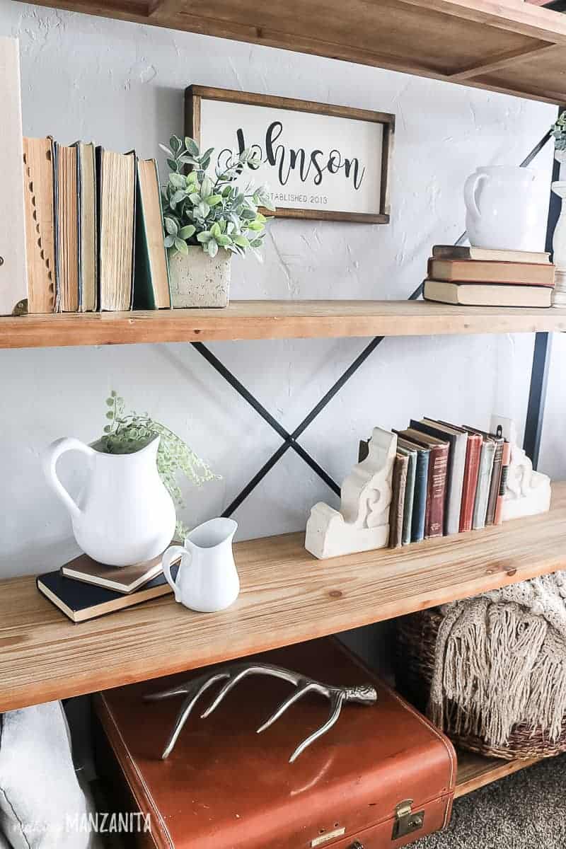 Vintage decor displayed on a shelving unit with farmhouse style showing white farmhouse jugs, vintage books, corbles, greenery and faux plants, farmhouse sign, vintage suite case and silver antler