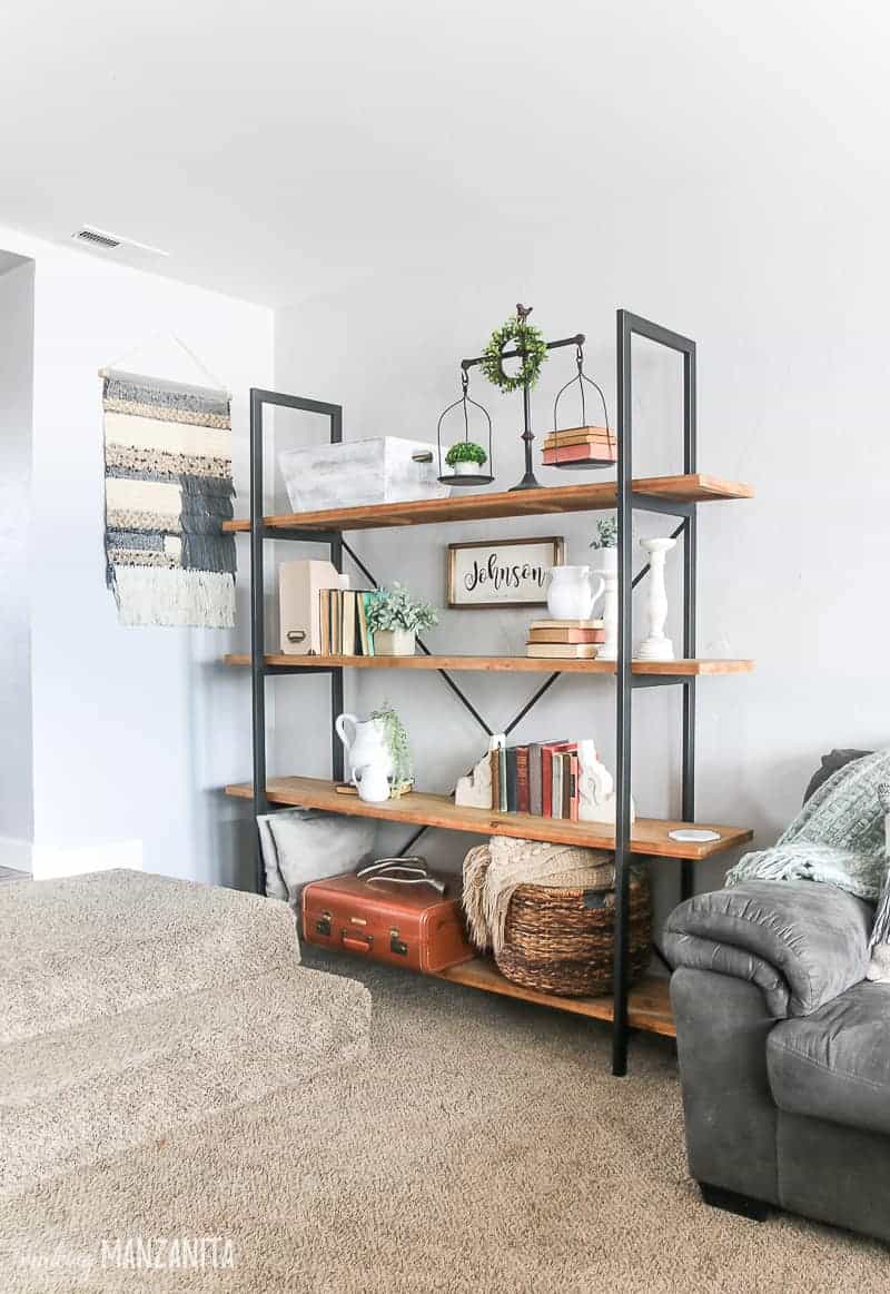 Corner of living room with large bookcase with rustic wood shelves, metal brackets, farmhouse decor on shelves, light gray paint on walls and woven wall hanging 