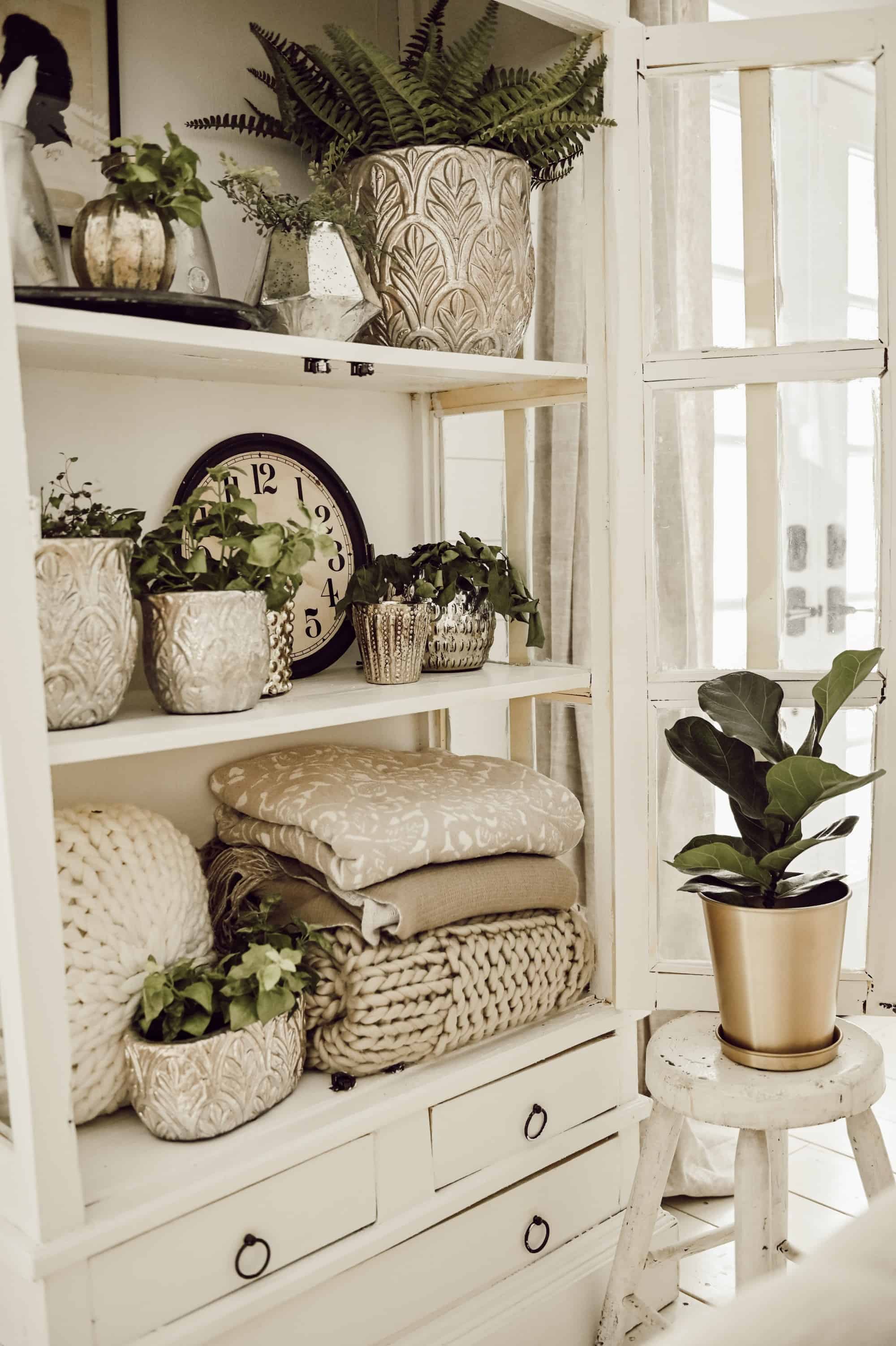 White vintage cabinet with doors open showing farmhouse styled decor like plants, folded cozy planets, clock