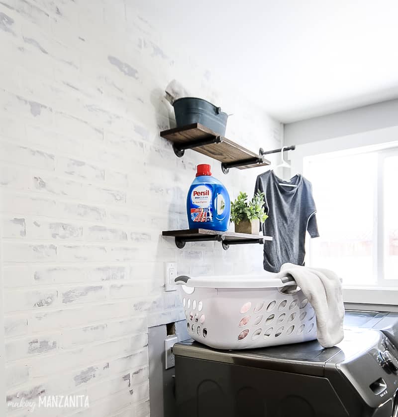 Farmhouse style laundry room with faux brick wall and industrial floating shelves above washer and dryer with bottle of Persil laundry detergent. There is a basket on top of the washer and dryer as well 