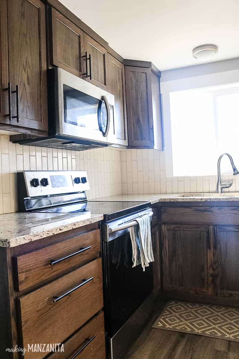 Dark wood kitchen cabinets with black modern hardware, stainless steel appliances and tan vertical backsplash with tan granite countertops and a window over the sink