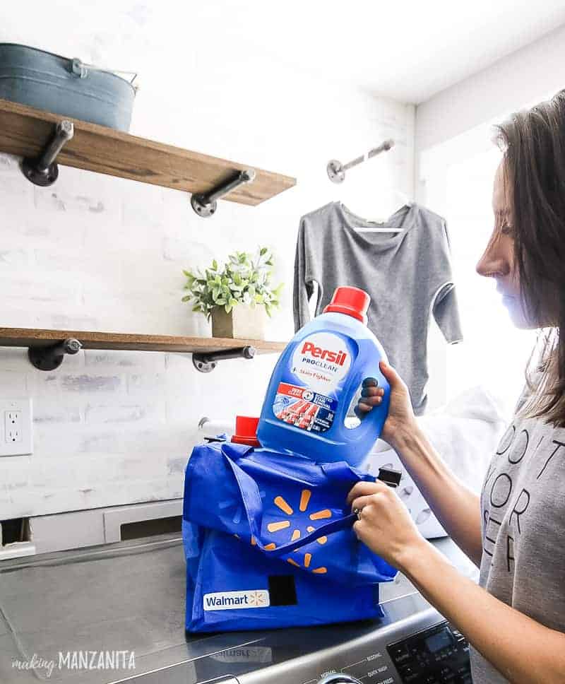 Woman taking a bottle of Persil laundry detergent out of a blue Walmart bag, which is sitting on top of washer and dryer with farmhouse style laundry room shown in the background