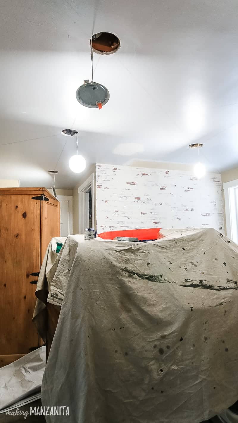 Renovations in progress in a laundry room with a drop cloth laid over washer & dryer in the middle of the room, recessed lights hanging out of the ceiling, faux brick wall shown in background