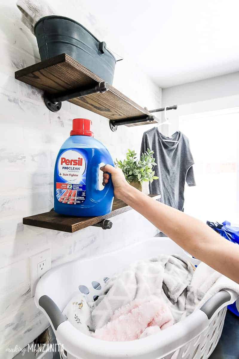 Woman's hand reaching for a bottle of Persil laundry detergent on a shelf in a farmhouse style laundry room over a laundry basket full of clothes sitting on top of the washing machine