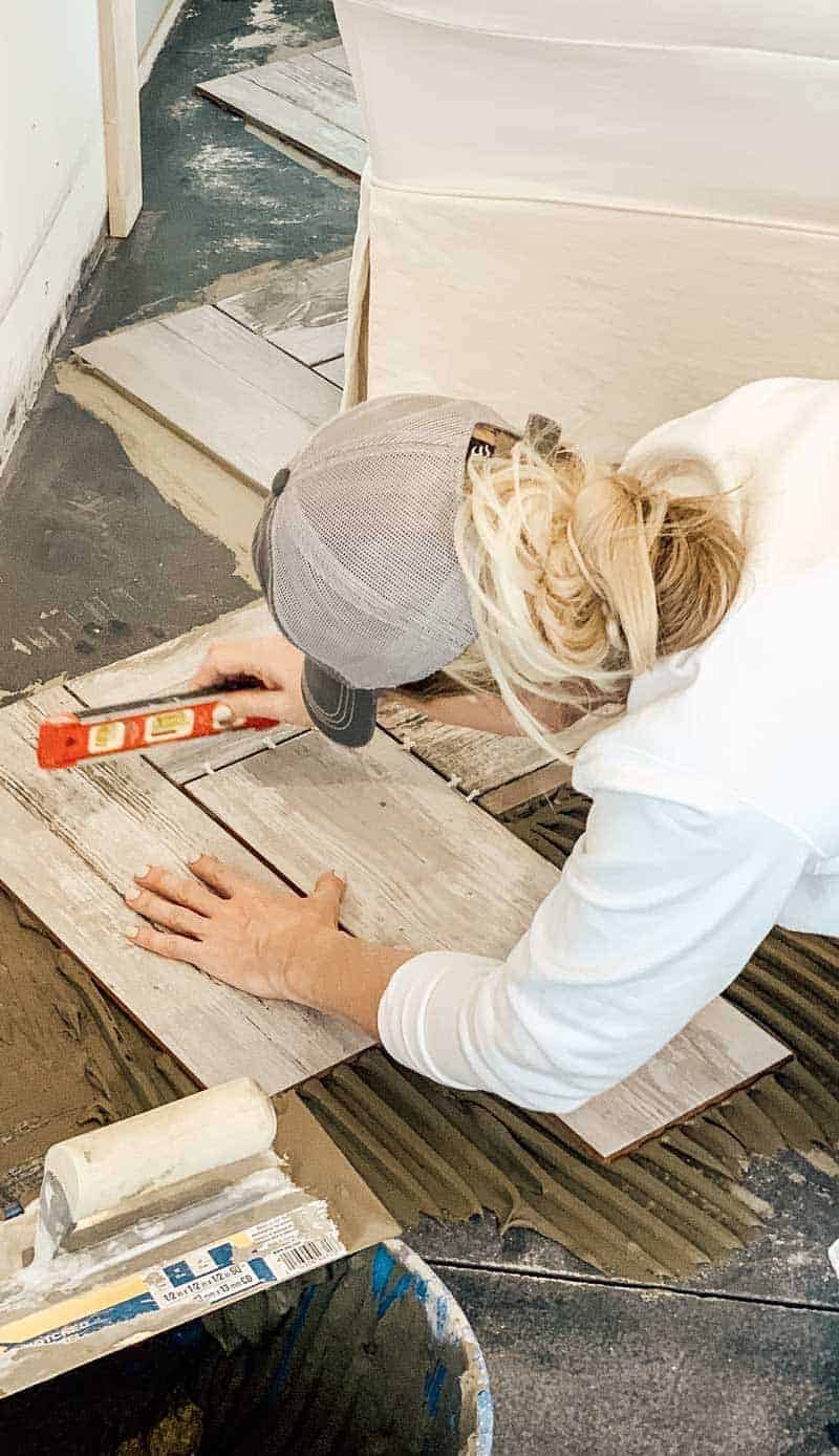 Woman installing herringbone pattern tile flooring holding a level and wearing a baseball cap