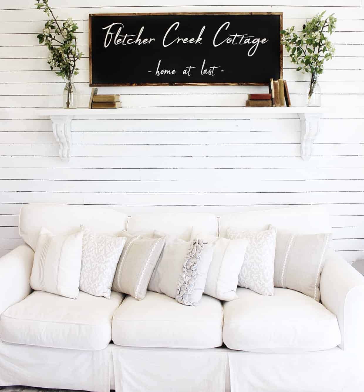 Farmhouse style living room with white couch and neutral throw pillows with white skinny shiplap walls, a shelf above the couch with a farmhouse style sign that says Fletcher Creek Cottage home at last and vintage books and greenery