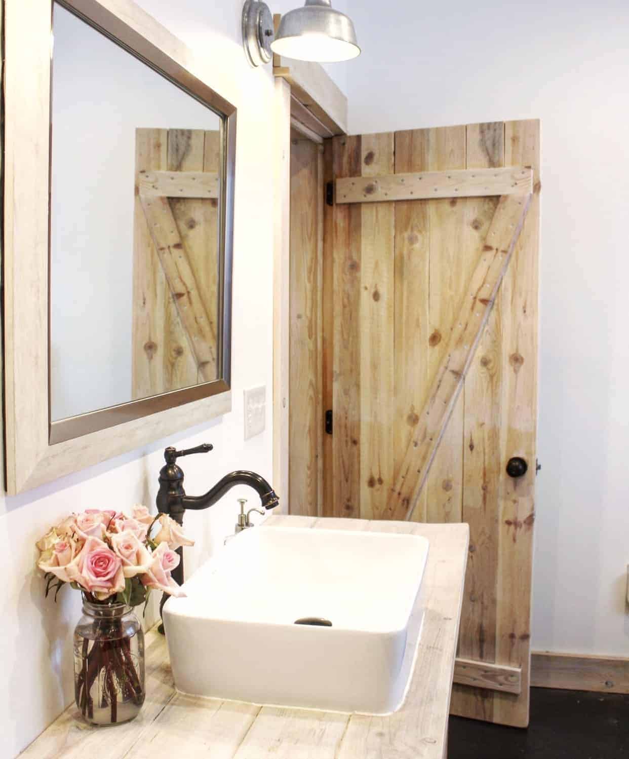 Farmhouse style bathroom with a wooden barn door, pendant barn style lighting, large farmhouse basin sink above wooden countertops with a vase of pink roses on the counter