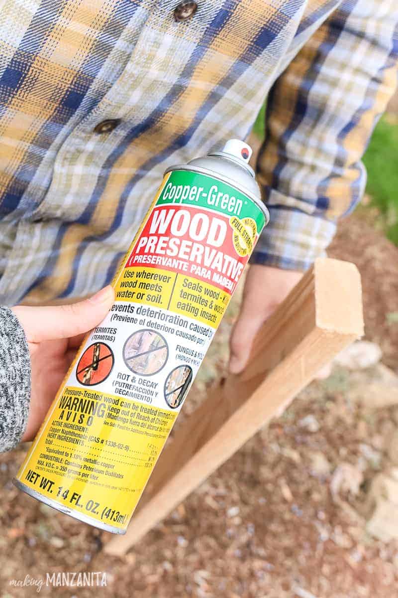 Bottle of spray copper green wood preservative held in front of a piece of 2x4 