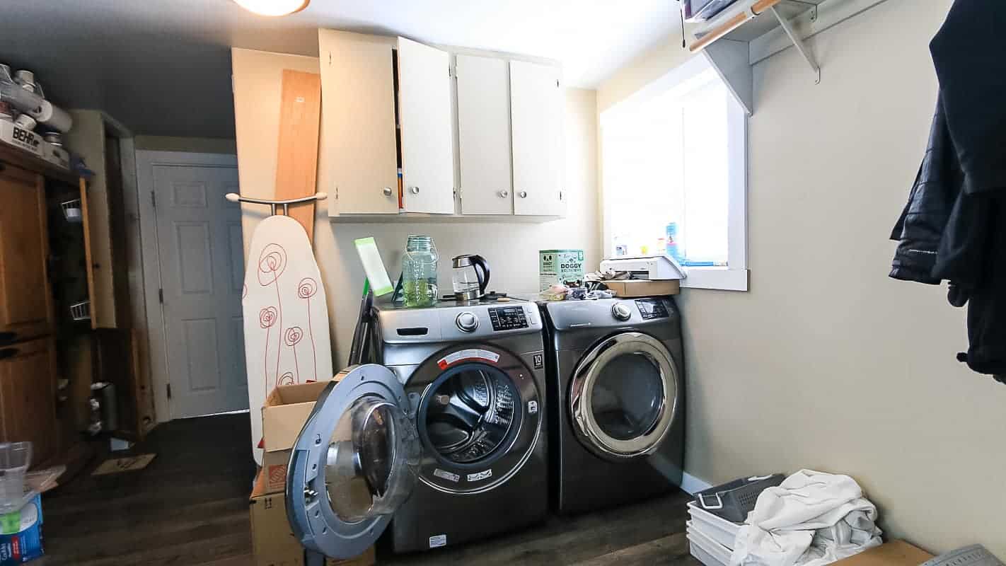 Messy laundry room with tan walls, side by side washer and dryer, upper cabinet, window and piles of clutter everywhere