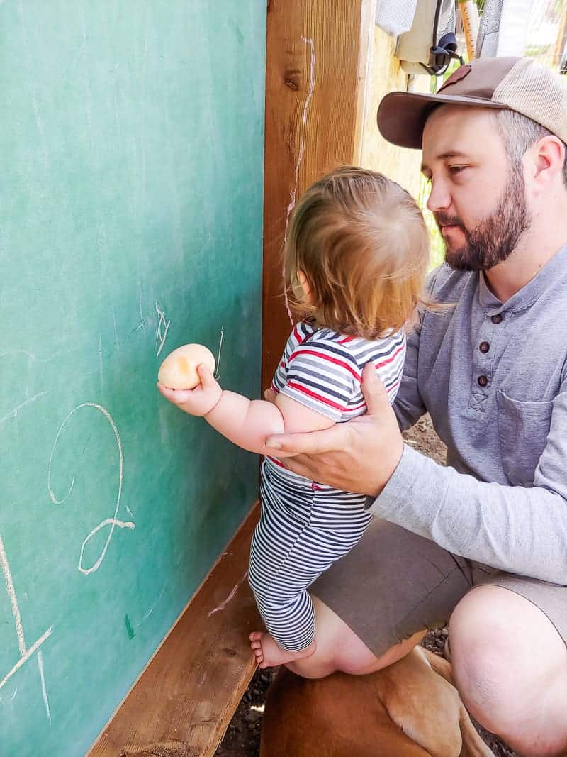 Logan helping out baby girl draw with a chalk egg on the large hanging outdoor chalkboard.