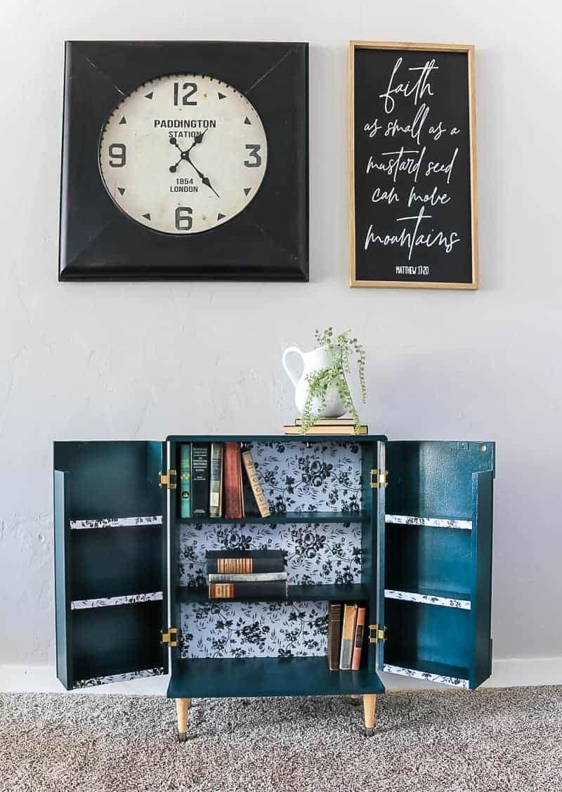 Full image of the opened dark green boho style laminate cabinet makeover showing the inside with books, clock and frame above it and farmhouse decor on top.