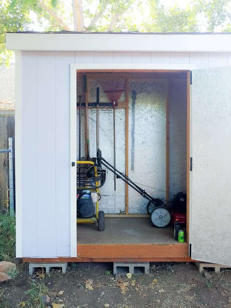 Opened door of garden tool storage showing gardening tools hanging on wall mounted hooks, lawn mower and other gardening machines.