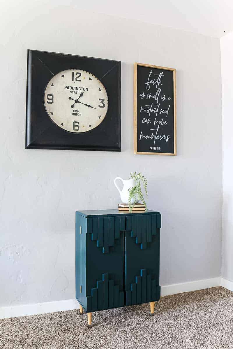 Slight side view of dark green boho laminate cabinet makeover with industrial large clock and framed quote sign above it with white pitcher and vintage books on top