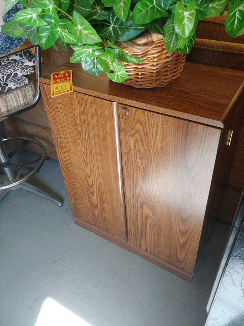 Old Media VHS storage cabinet in a thrift store with a sale tag of  on it with a basket of artificial plant above before photo of laminate cabinet makeover