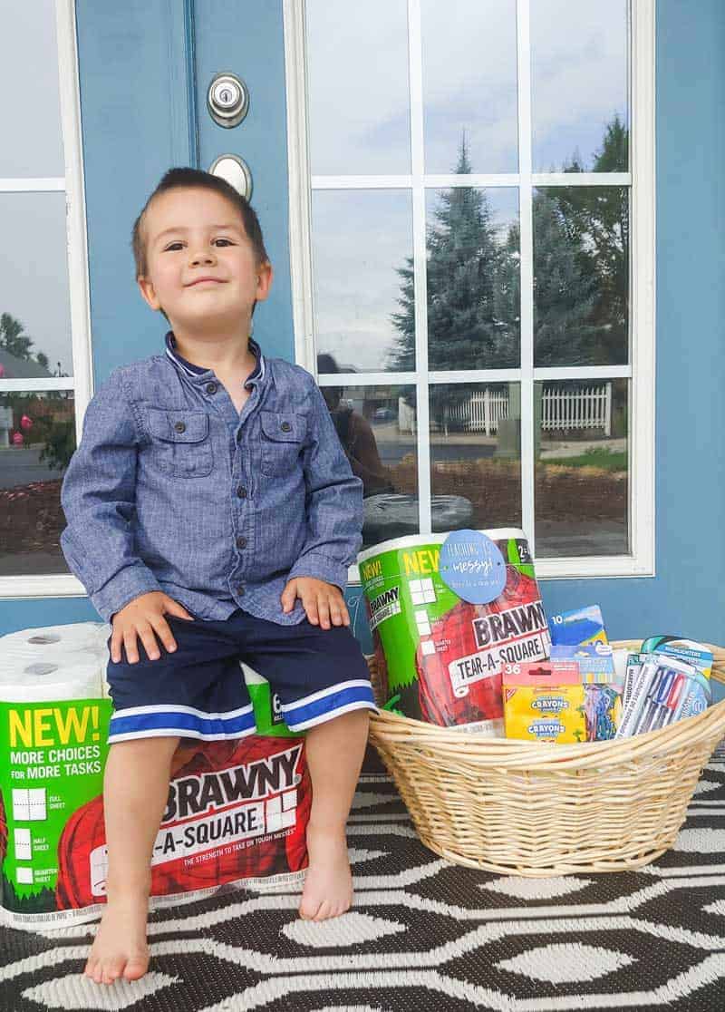 Preschooler boy sitting on the paper towel pack and a teacher gift set basket beside him