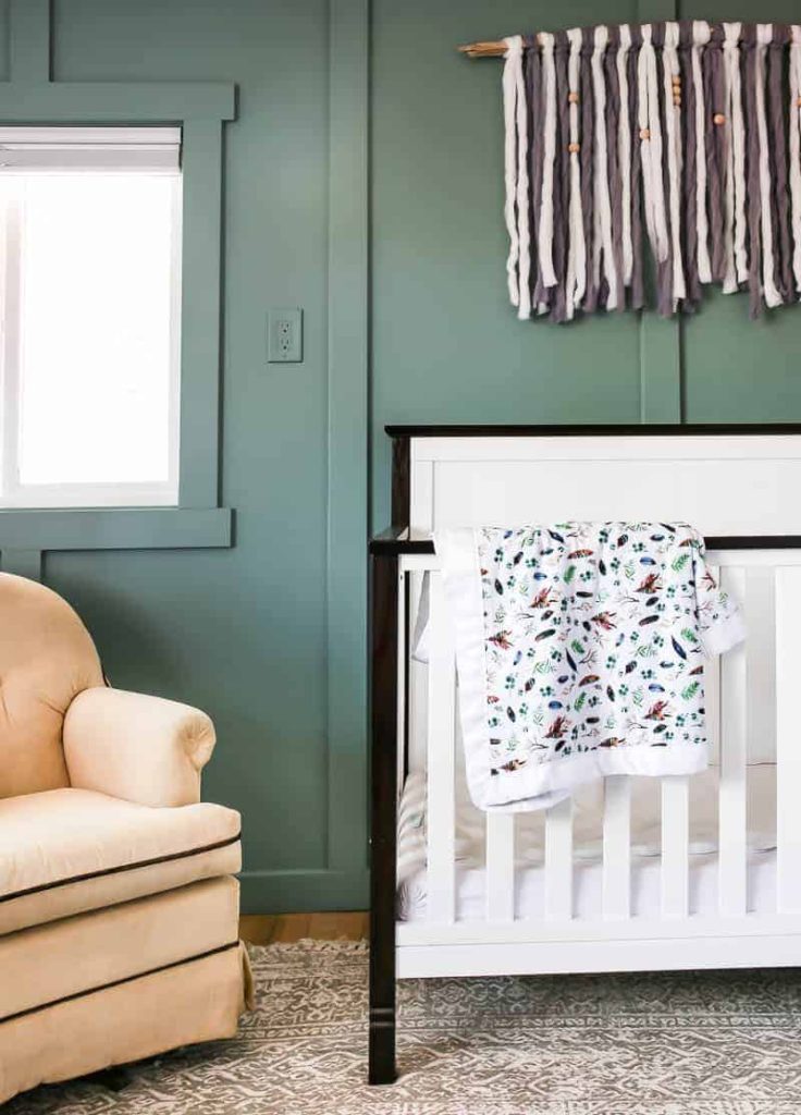 White farmhouse style crib with black accents with a blanket laying over the edge against a green board and batten wall with boho wall hanging above it with a vintage inspired rug on floor and tan chair sitting next to it in corner