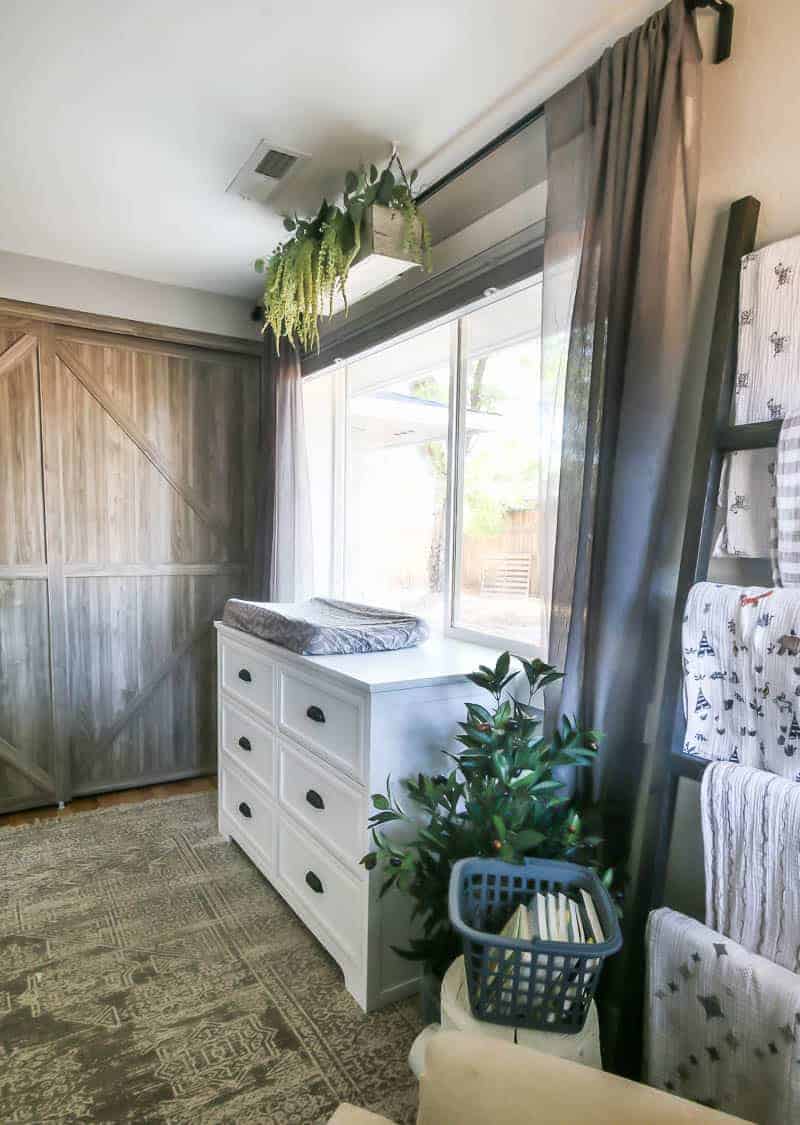 The changing table in our baby girl's nursery sits in front of a large window. Next to the changing table the closet, complete with farmhouse style barn doors, and a nursing area decorated with plants and a baby blanket ladder.