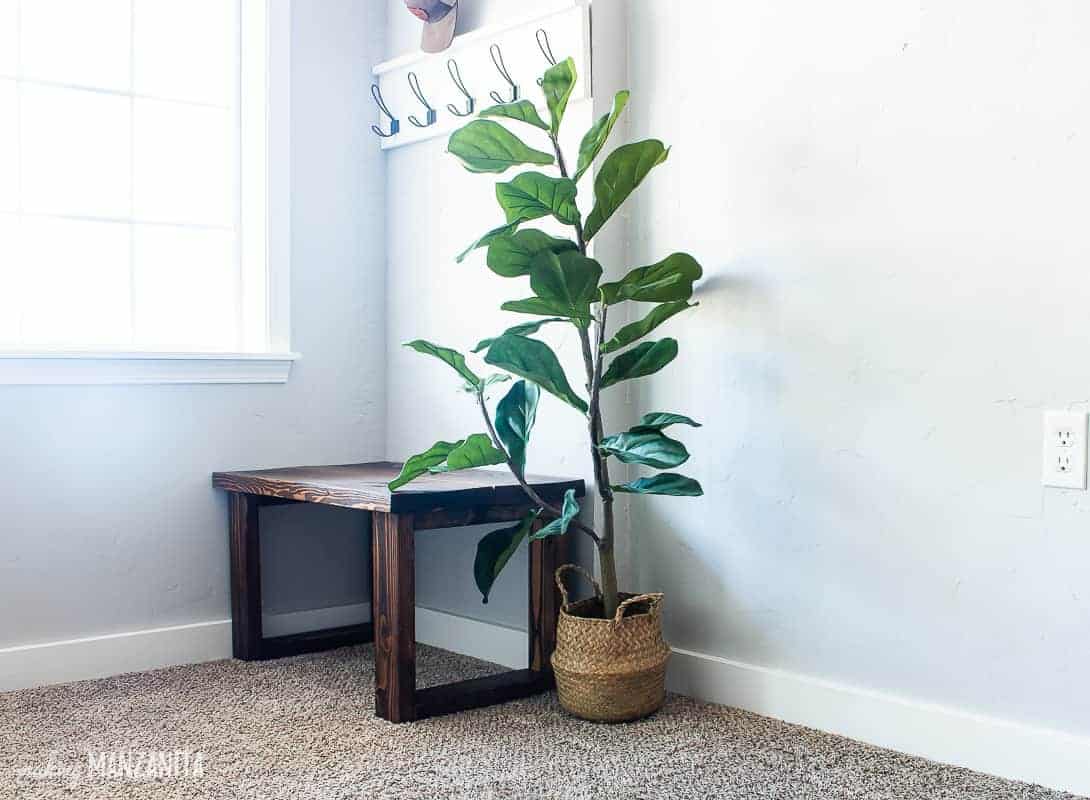 Side shot of the dark wood bench in the small entryway in corner of living room with an faux fiddle leaf fig tree next to in with window in background