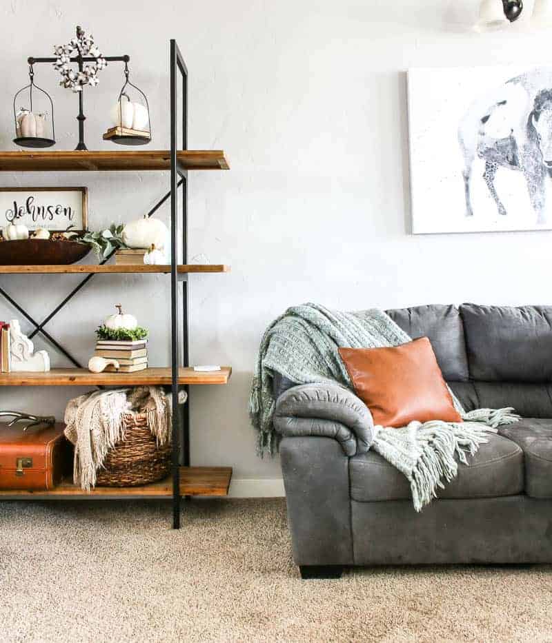 Our Fall themed living room bookshelf, adorned with my favorite living room shelf decor, next to our sectional sofa with a neutral throw blanket and rust colored throw pillow.