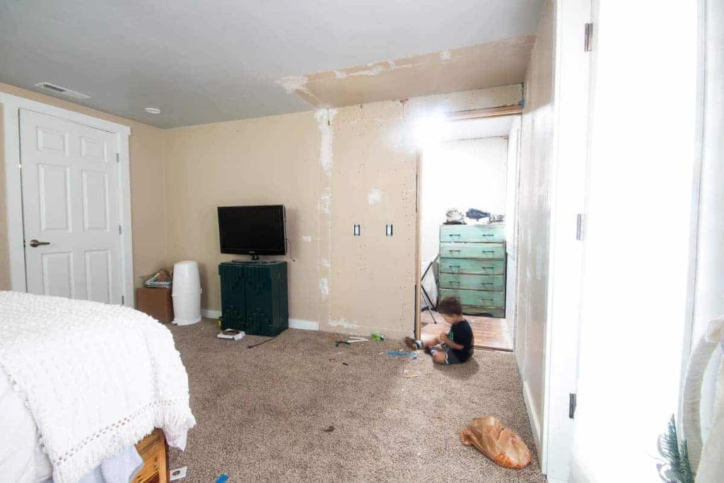 An in progress photo of the renovations of the master bedroom closet. Walls have been removed, leaving a large open space and construction marks on the wall. A young boy sits on the carpet where the closet walls used to be.